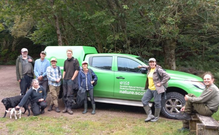 8 smiling volunteers in a woodland standing by a Scottish Natural Heritage van with two dogs.