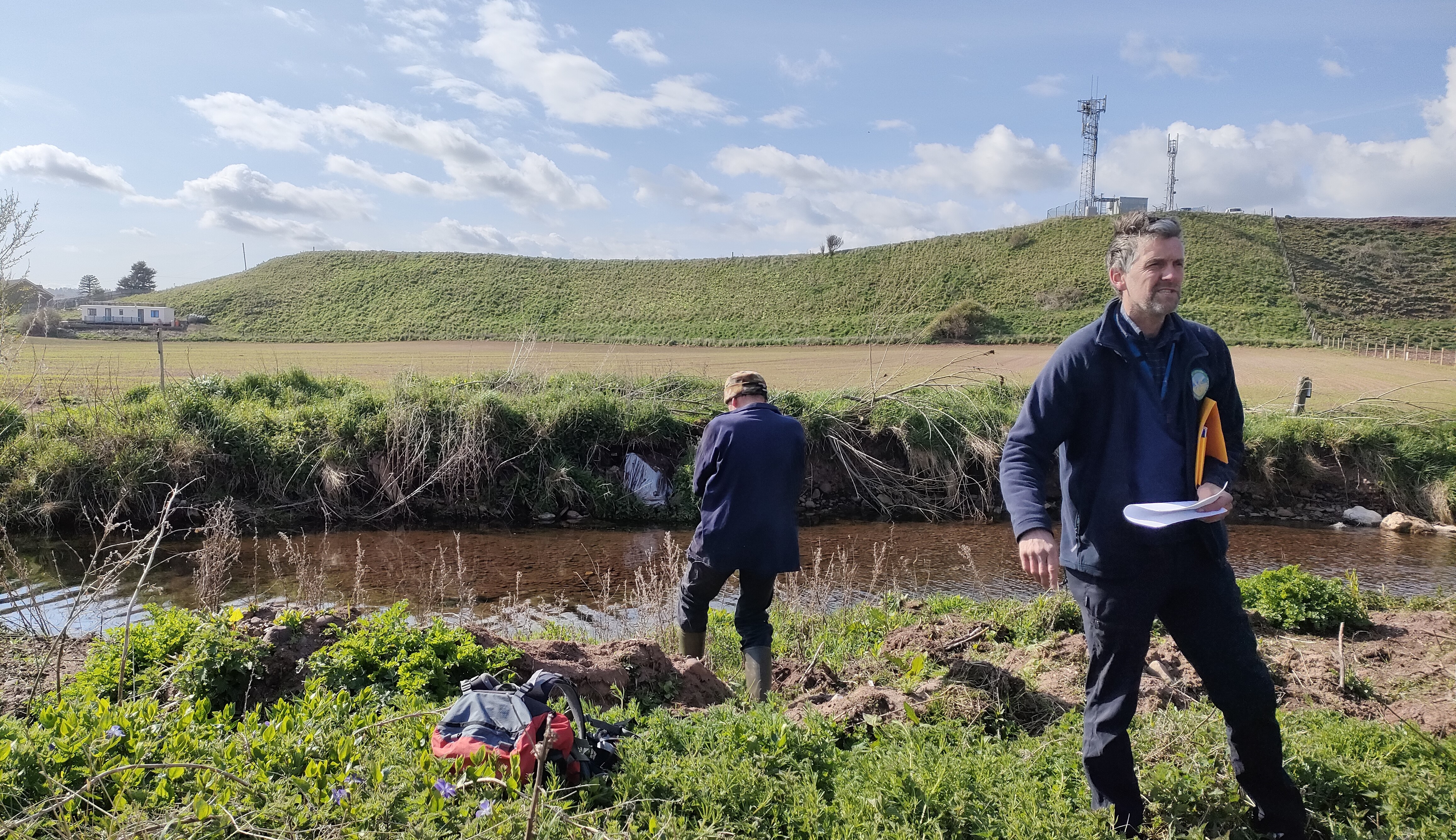 A small river with an eroded riverbank. It is sunny and there are two men near the river.